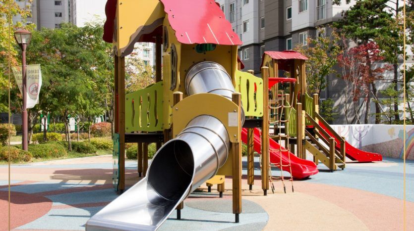 Une aire de jeux colorée avec toboggan et structures d'escalade, installée sur un tapis en caoutchouc, dans un parc urbain entouré de bâtiments et d'arbres par une journée ensoleillée à Dubaï.