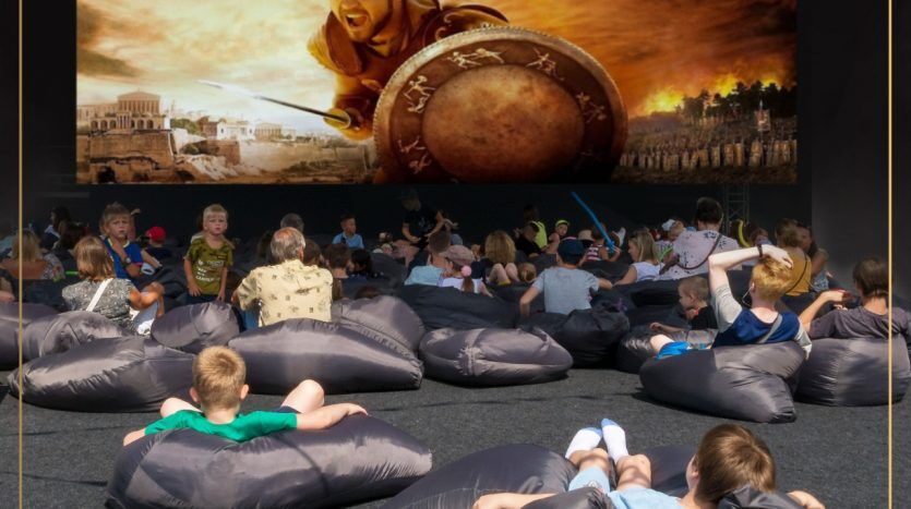 Des enfants assis sur des poufs dans une zone d'événement public avec une grande toile de fond montrant une scène pleine d'action d'un guerrier avec un bouclier et une lance, avec des publicités pour investir à Dubaï visibles sur des banderoles entourant la zone.