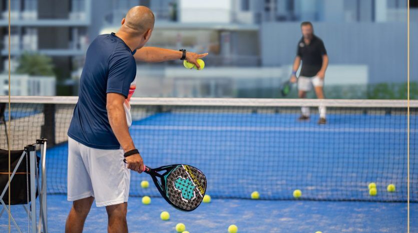 Un homme en chemise bleue jouant au padel sert tandis qu'un autre homme attend le retour sur un terrain extérieur près d'une villa de Dubaï, rempli de balles jaunes éparpillées.