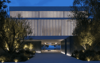 Bâtiment moderne de Dubaï éclairé au crépuscule avec un design symétrique comportant une passerelle centrale menant sous une grande structure surélevée flanquée de rangées d'arbres.