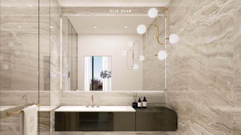 Une salle de bains luxueuse dans une villa à Dubaï avec des murs et des sols en marbre, avec un grand miroir arborant le logo « Elie Saab ». En dessous, il y a une élégante vanité noire avec des robinets dorés.