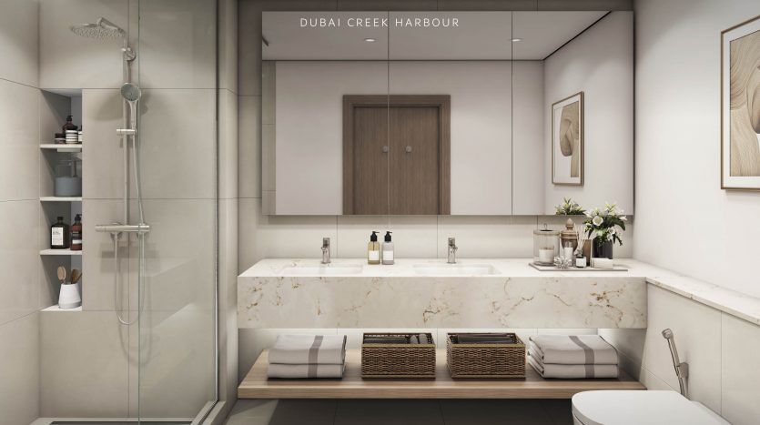 Intérieur de salle de bains moderne avec comptoir en marbre, double vasque, grand miroir, douche, toilettes et éléments décoratifs ; labellisée « la villa Dubai Creek Harbour – Emaar ».
