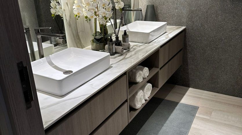 A modern bathroom featuring a wooden vanity with a white sink, marble walls and a vase of white flowers. A mirror, towels and toiletries are neatly arranged throughout the Dubai villa.