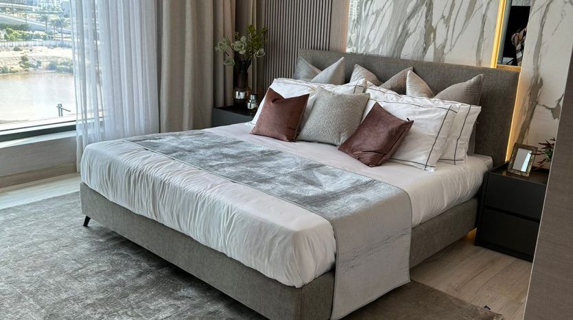 A modern room in a Dubai apartment featuring a king-size bed with beige and gray bedding, fluffy pillows, a marble wall and a view of the waterfront through large windows. The decor includes a round ceiling light and