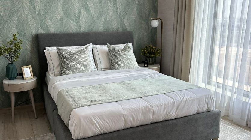 A modern bedroom in a Dubai apartment featuring a gray upholstered bed with white bedding, geometric green wallpaper, sheer curtains and minimalist decor including a side table and plants.