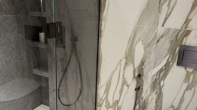 A modern bathroom with marble walls featuring large gray veins, a glass shower cubicle, built-in shelving and a white toilet next to a gray textured wall in an investment property in Dubai