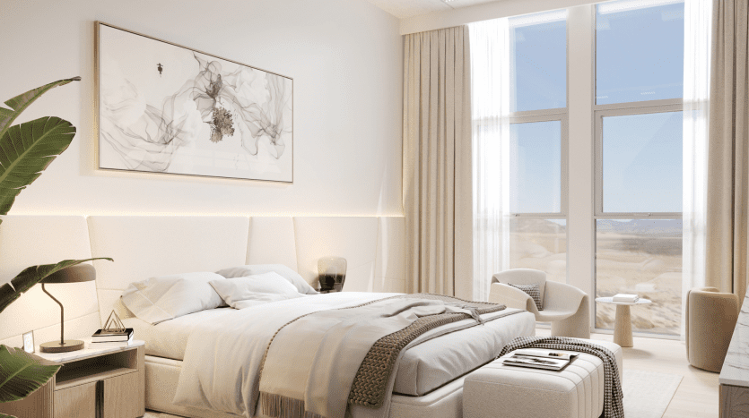 A bright, modern room in a Dubai apartment, with a king-size bed covered in neutral-toned bedding, flanked by two small side tables. Large windows with sheer curtains look out onto a sandy beach, with