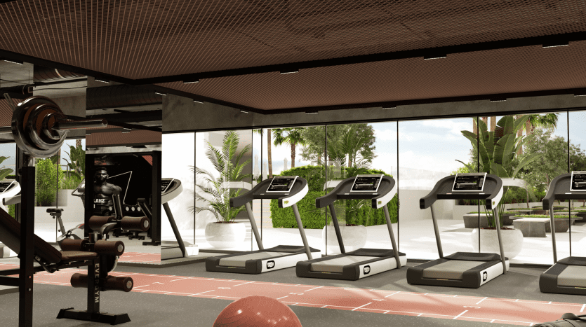 Modern gym interior with equipment including treadmills, weight machines and free weights. Large windows overlook the lush greenery typical of a Dubai villa.