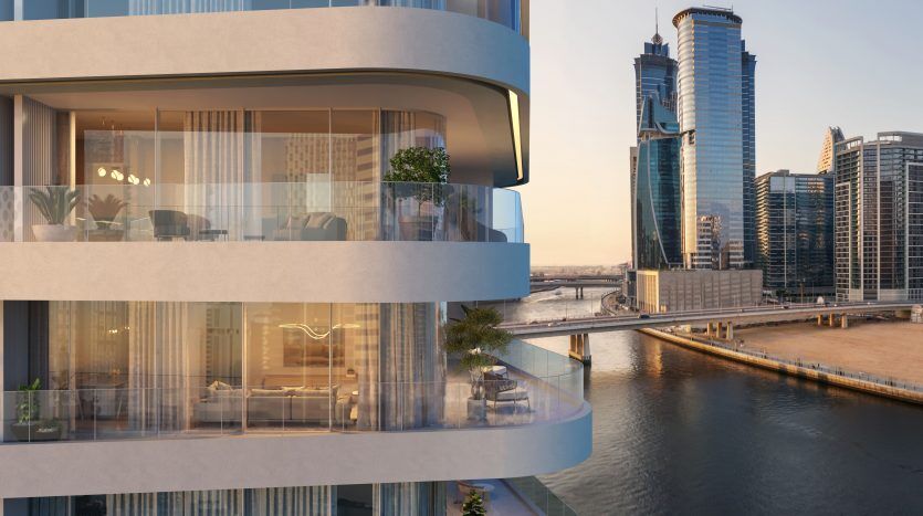 Vue extérieure d'un bâtiment moderne à plusieurs étages avec des balcons incurvés, des intérieurs bien éclairés, sur un paysage urbain avec des tours de grande hauteur et une rivière au coucher du soleil à Dubaï.
