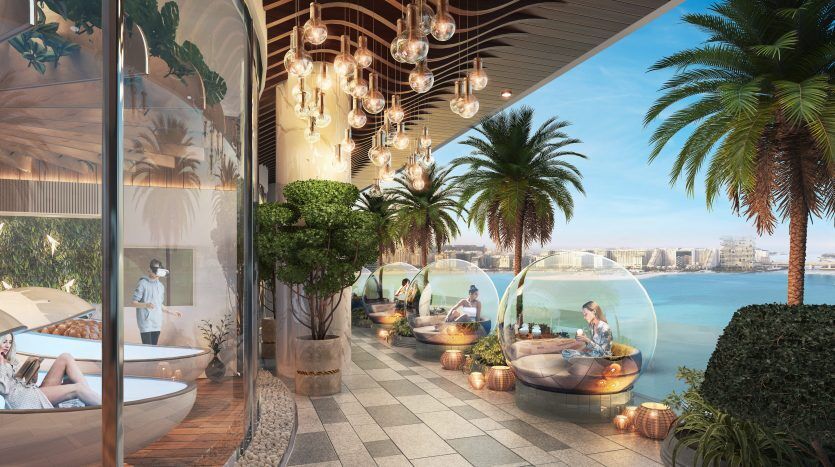 Luxueux salon extérieur au bord de l'eau avec d'élégantes suspensions, des plantes luxuriantes et des capsules à bulles transparentes contenant des coins repas, le tout donnant sur une vue panoramique d'une villa à Dubaï.