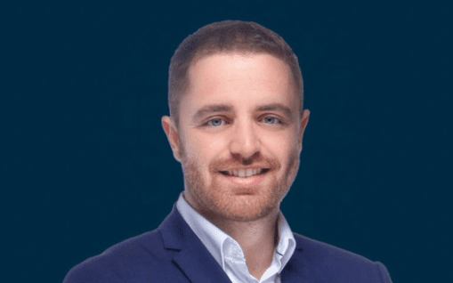 Un portrait professionnel d'un homme souriant avec une barbe, vêtu d'un blazer bleu marine et d'une chemise blanche, sur un fond bleu sur le thème "immobilier Dubaï".