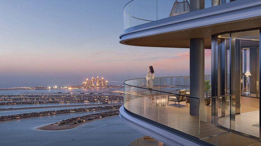 Une femme se tient sur le balcon moderne d’une villa de Dubaï surplombant un paysage urbain côtier au crépuscule, avec des gratte-ciel illuminés et une mer calme en arrière-plan.