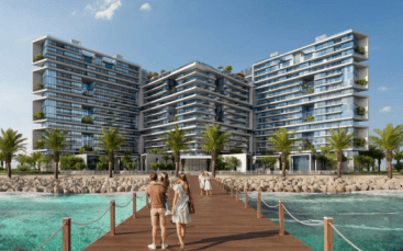 Bâtiments résidentiels modernes à plusieurs niveaux en bord de mer avec une verdure luxuriante sur les balcons sous un ciel bleu clair. Une jetée en bois avec des personnes marchant vers le complexe est visible au premier plan. Idéal pour immobilier Dubaï