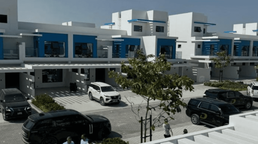 A modern residential area of Dubai with contemporary white and blue houses, each with flat roofs and balconies. The street is lined with parked cars and a few passers-by. Trees add greenery to the