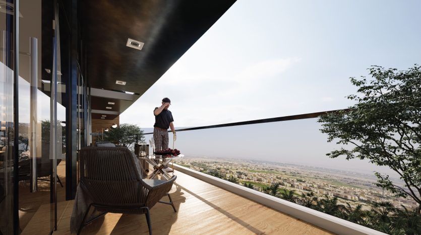 Une personne se tient sur un balcon donnant sur un vaste panorama et prend une photo avec son téléphone. Le balcon, un endroit idéal pour l'immobilier à Dubaï, présente un mobilier moderne et une verdure luxuriante au loin.