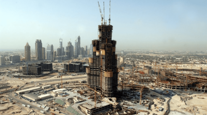 Vue grand angle d'un grand chantier de construction à Dubaï avec la structure squelettique d'un immeuble imposant au premier plan, entouré de divers autres immeubles de grande hauteur en construction, sous un ciel brumeux.