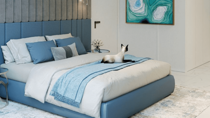 Une chambre moderne dans un appartement de Dubaï comprenant un grand lit bleu avec une tête de lit moelleuse, une literie douce bleue et grise et un chat siamois assis au pied du lit. Une peinture vibrante de vert et de bleu est accrochée au mur.