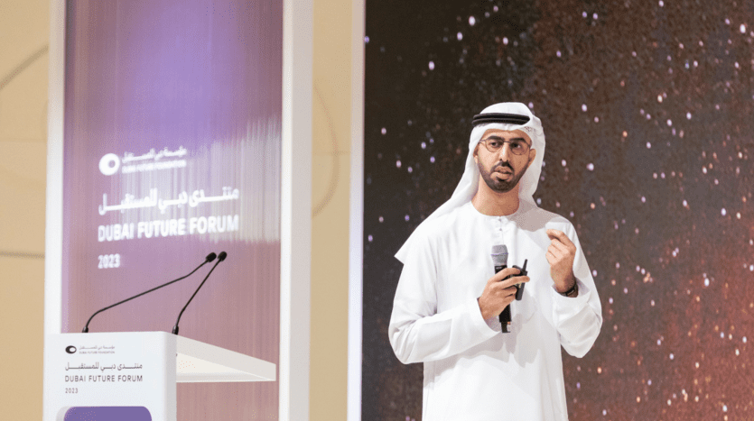 A man in traditional Emirati dress speaking on a podium with a microphone at the Dubai Future Forum 2023, with a digital display featuring Dubai real estate in the background.