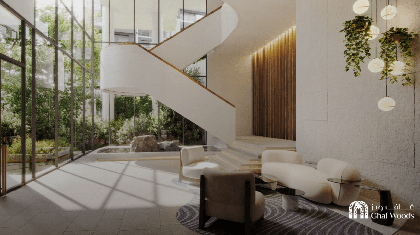 L'image montre un atrium moderne et ensoleillé avec de grandes fenêtres et un escalier en colimaçon blanc. L'espace est décoré avec un mobilier contemporain comprenant des chaises blanches et beiges, une table basse et une plante en pot. Un logo pour « Ghaf Woods » est affiché bien en évidence dans le coin inférieur droit.