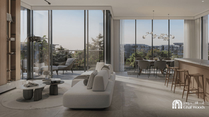 Un salon et salle à manger moderne à aire ouverte comprend un élégant canapé blanc, une table basse minimaliste, des baies vitrées, un lustre contemporain et un comptoir de bar avec des tabourets. À l’extérieur, le grand balcon du Ghaf Woods surplombe un paysage verdoyant.