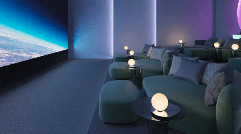 A modern, luxurious home theater with recliners and soft green poufs. Each seating area has a small table with a round lamp. A large screen displaying an image of space with the Earth visible dominates the wall opposite the sitting area.