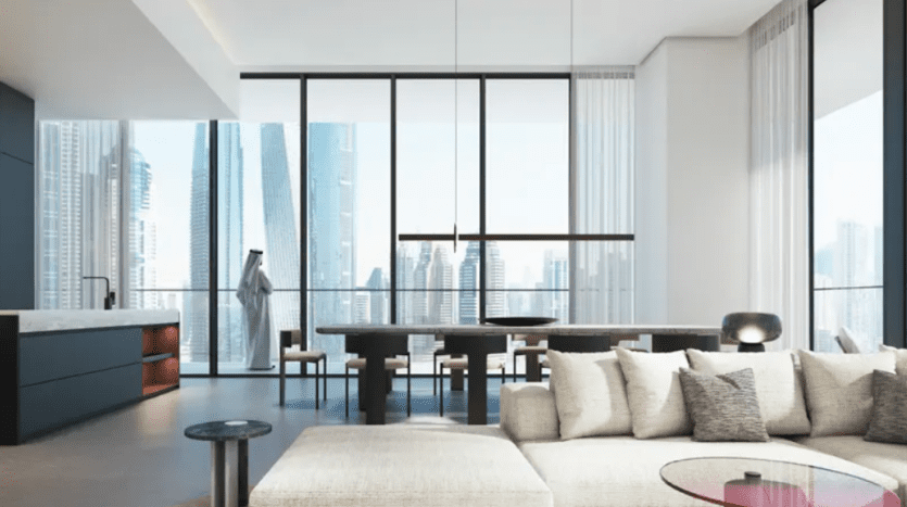 A modern living and dining room with bay windows highlighting a skyscraper-filled cityscape. The space features minimalist furnishings, including a white sofa, a black dining table with chairs and a kitchen island. One person stands by the window.