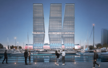 Una moderna scena sull'acqua al tramonto è caratterizzata da due grandi edifici identici con linee orizzontali che formano una forma a V. Le persone socializzano lungo la passeggiata, con gli yacht ormeggiati nel porto turistico in primo piano. L'area è illuminata da luci ambientali.