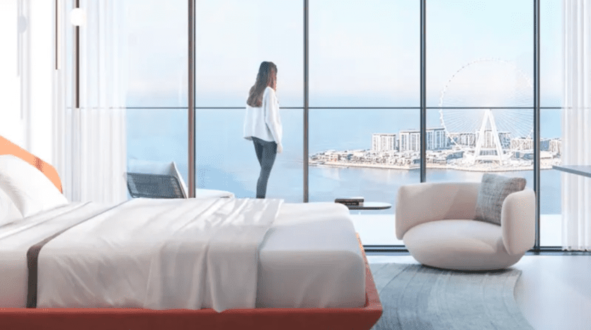 A long-haired person stands in front of the picture windows of a bright, modern room with a king-size bed, armchair and blue carpet. The view includes a distant Ferris wheel and a vast expanse of water, suggesting a high-rise building near a waterfront.