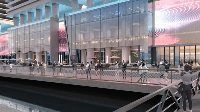 View of the waterfront from a modern glass building with large digital screens displaying abstract motifs. People walk along the promenade, some stopping to take photos or chat. The sky is clear and the general atmosphere is lively and urban.