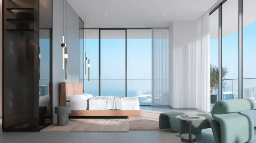 A modern room with bay windows offering ocean views. The room features a low bed, a small side table and green armchairs. Minimalist hanging lights hang from the ceiling and bright curtains frame the windows, letting in natural light.