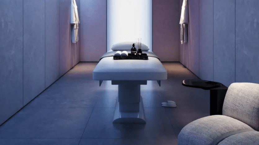 A minimalist, dimly lit spa room includes a massage table with towels, essential oils and aromatherapy items neatly arranged. The walls are smooth and gray and a soft chair sits in the corner, emphasizing a tranquil atmosphere.