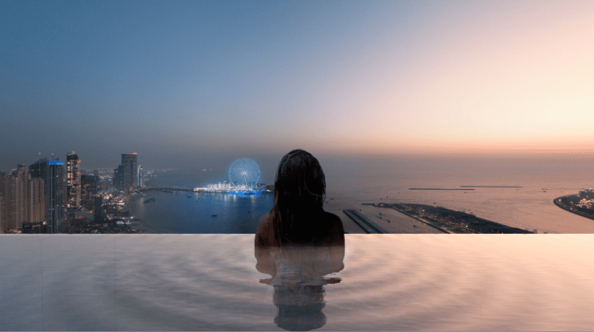A person is shown from behind, sitting in an infinity pool at dusk at the W Residences Dubai Harbour. The view looks out over the rooftops of the city, with tall buildings, an illuminated Ferris wheel and a calm sea under a sky gradated from blue to pink. Ripples surround the person in the water.
