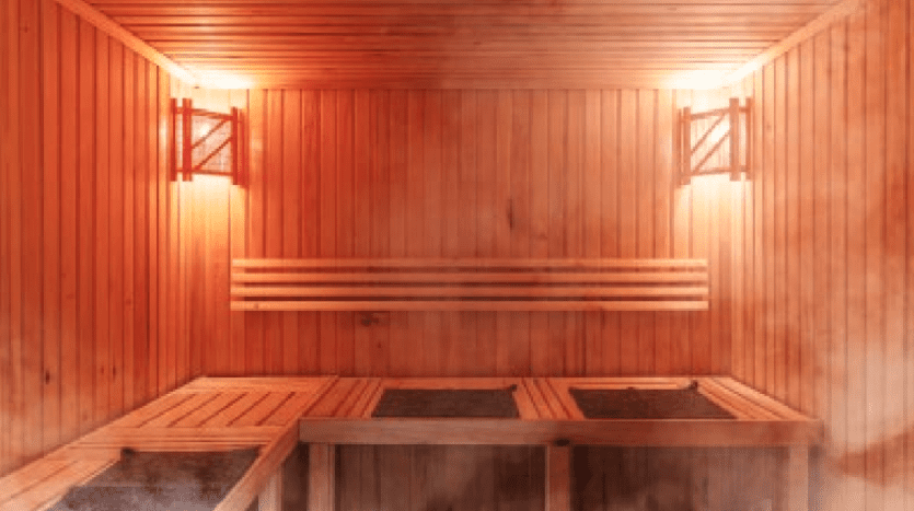 A wooden sauna at W Residences Dubai Harbour features warm, bright lights on the walls and steam rising from the floor. Wooden benches line the walls beneath a wooden slatted ceiling. The ambience is warm and welcoming, perfect for relaxing in luxury.