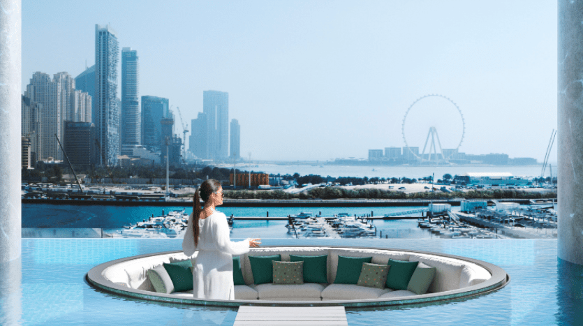 A woman in a white dress stands in a raised, circular, sunken seating area surrounded by water in the luxurious port of W Residences Dubai, overlooking a marina with boats and the city's skyline. A Ferris wheel can be seen in the distance against a clear blue sky.