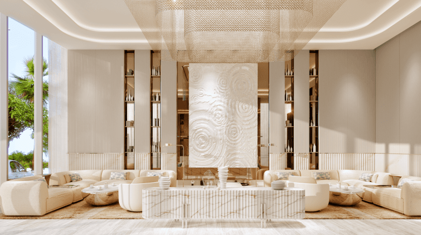 A luxurious lounge with high ceilings, with beige and cream decor reminiscent of The Pier. It features two large cream-colored sectional sofas, several round coffee tables and a large, intricate central artwork. Large windows let in plenty of natural light, and greenery is visible outside.