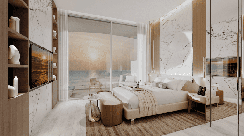 Modern hotel room with marble accents, featuring a king-size bed, wall-mounted TV and ornate built-in shelving. A sliding glass door reveals a balcony with seating, overlooking the jetty and its serene view of the ocean at sunset. Neutral tones and elegant furnishings create a luxurious atmosphere.