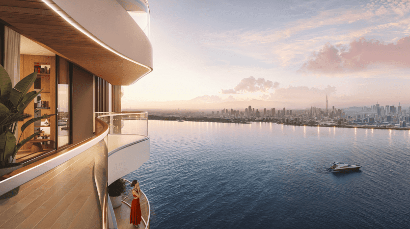 A person dressed in a red dress stands on a curved balcony at The Pier, overlooking a tranquil body of water at sunset. The balcony is part of a modern building with wood and glass elements. In the distance, a city skyline and a yacht are visible on the horizon.