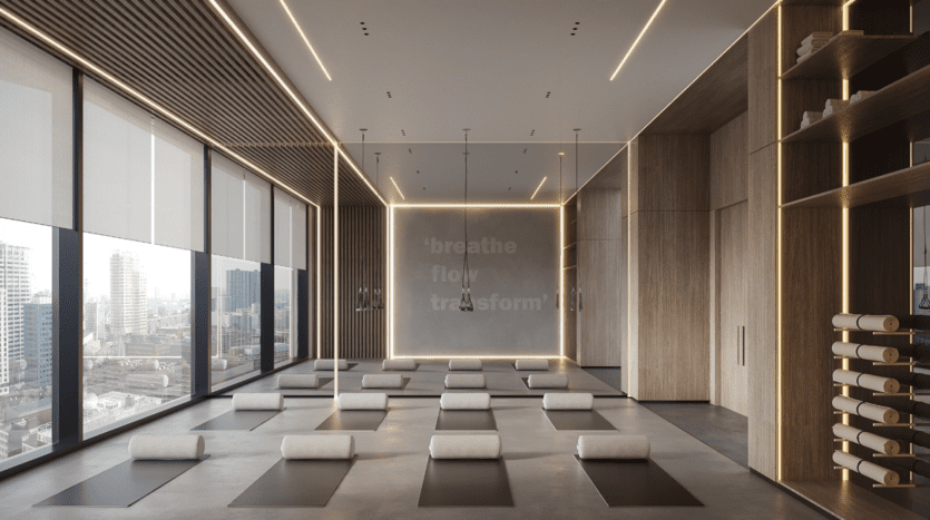Un studio de yoga moderne avec des tapis roulés et des coussins au sol, des touches de bois et de grandes fenêtres offrant une vue sur la ville. Le mur affiche les mots « respirer, couler, transformer » illuminés par un éclairage doux.