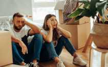 Un hombre y una mujer sentados en el suelo, con aspecto cansado y estresado, rodeados de cajas de mudanza en una habitación soleada. Mientras reflexionan sobre cómo amueblar un piso en Dubai, una gran maceta destaca en primer plano, ofreciendo una pizca de serenidad en medio del caos.
