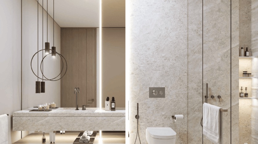 La salle de bains moderne du Safa Gate Damac est dotée d'une bande lumineuse verticale qui divise l'espace. À gauche, une double vasque avec des suspensions et des produits de toilette complète le design minimaliste à la texture de pierre, tandis qu'à droite, des toilettes et une douche vitrée complètent cet élégant sanctuaire.
