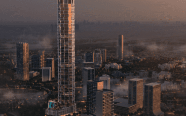 Un grand gratte-ciel moderne domine un paysage urbain au crépuscule, entouré de plusieurs bâtiments plus petits et d'une végétation luxuriante. Des lumières illuminent les bâtiments et un ciel brumeux crée une scène tranquille et atmosphérique.