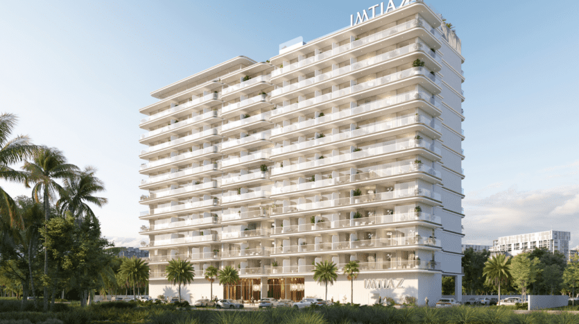 Un immeuble d'appartements moderne, blanc, de plusieurs étages - Cove Edition 6 de Imtiaz - avec de grands balcons et des palmiers à l'avant se dresse sous un ciel bleu limpide. L'enseigne "IMTIAZ" couronne le sommet et apparaît au-dessus de l'entrée principale.
