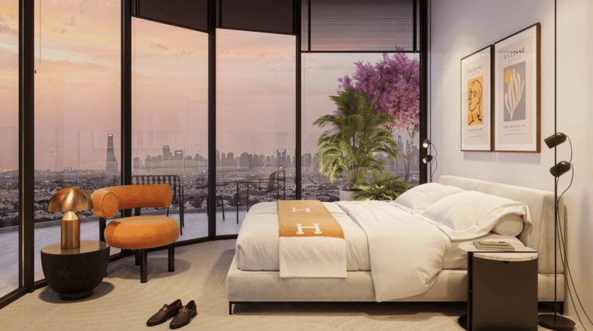 Chambre à coucher moderne avec des fenêtres allant du sol au plafond et donnant sur une ville au coucher du soleil, un lit en peluche avec une couverture beige, une chaise orange, des tables de chevet, des œuvres d'art encadrées au mur, une plante en pot et des chaussures sur le sol.