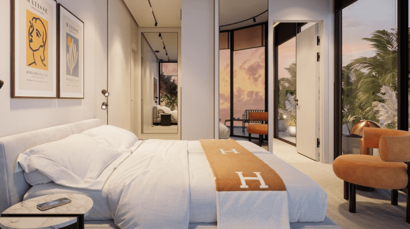 Chambre à coucher moderne avec un lit bien fait, une couverture beige et des oreillers blancs. De grandes fenêtres révèlent un coucher de soleil éclatant et de la verdure à l'extérieur. Le mobilier minimaliste comprend des chaises, une table d'appoint avec un téléphone et des œuvres d'art sur les murs.