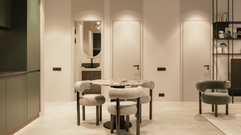 Salle à manger moderne et minimaliste avec une table ronde en marbre et quatre chaises rembourrées, sur des murs beiges. Il y a trois portes fermées, un coin cuisine vert sur la gauche et des étagères ouvertes sur la droite.