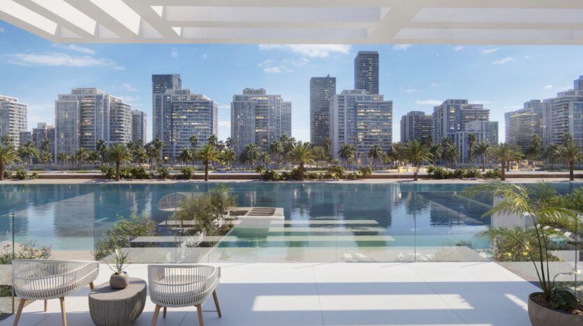 Profitez de la vue depuis un balcon moderne à Ayaan Heights 1 by IGO, avec deux chaises donnant sur une voie d'eau bleue claire, des palmiers se balançant, et une ligne d'horizon de grands bâtiments modernes sous un ciel bleu lumineux.