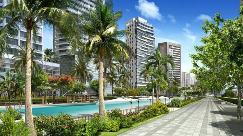 Une large allée bordée d'arbres longe une piscine et des espaces de détente à Ayaan Heights 1 by IGO, entouré d'une végétation luxuriante et de gratte-ciel modernes sous un ciel bleu lumineux.