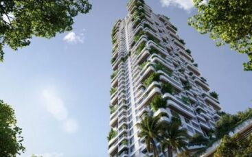 Un grand gratte-ciel moderne, Talea by Beyond, avec des balcons blancs ornés de plantes et d'arbres verdoyants, vus d'en bas sur fond de ciel bleu et encadrés par des branches feuillues.