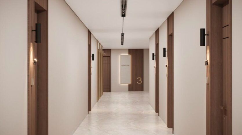 Un couloir moderne et propre au Marquis One Arjan présente des murs de couleur crème, des sols en marbre, des cadres de porte en bois et des appliques murales. Des portes bordent les deux côtés, et un panneau indiquant "3" est visible au bout du couloir.