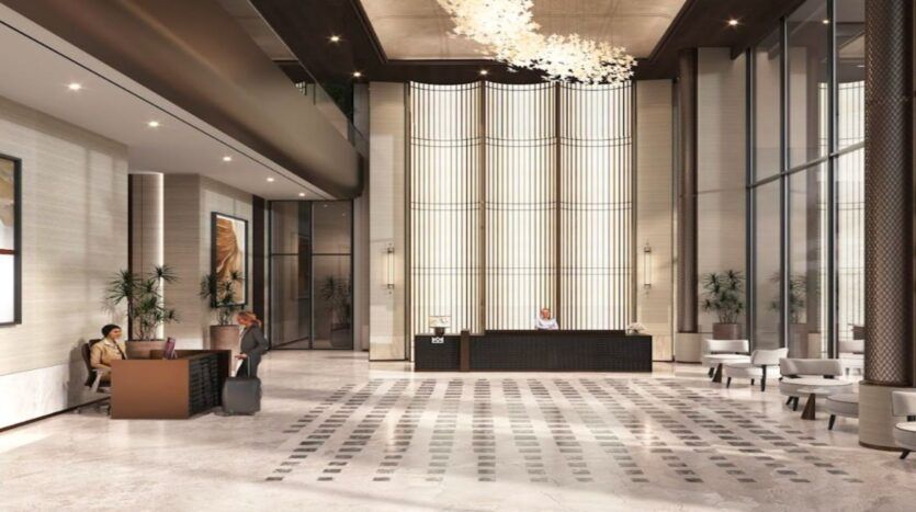 Hall d'entrée spacieux et moderne de l'hôtel Marquis One Arjan, avec de hauts plafonds, un éclairage élégant, un bureau de réception central, un coin salon à gauche, de grandes fenêtres, et deux personnes interagissant près des bagages.